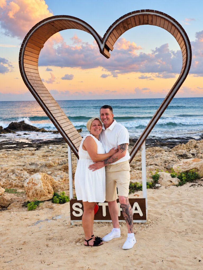 Alex and Paul embracing beneath the wooden heart at Sirens Beach after their sand blessing